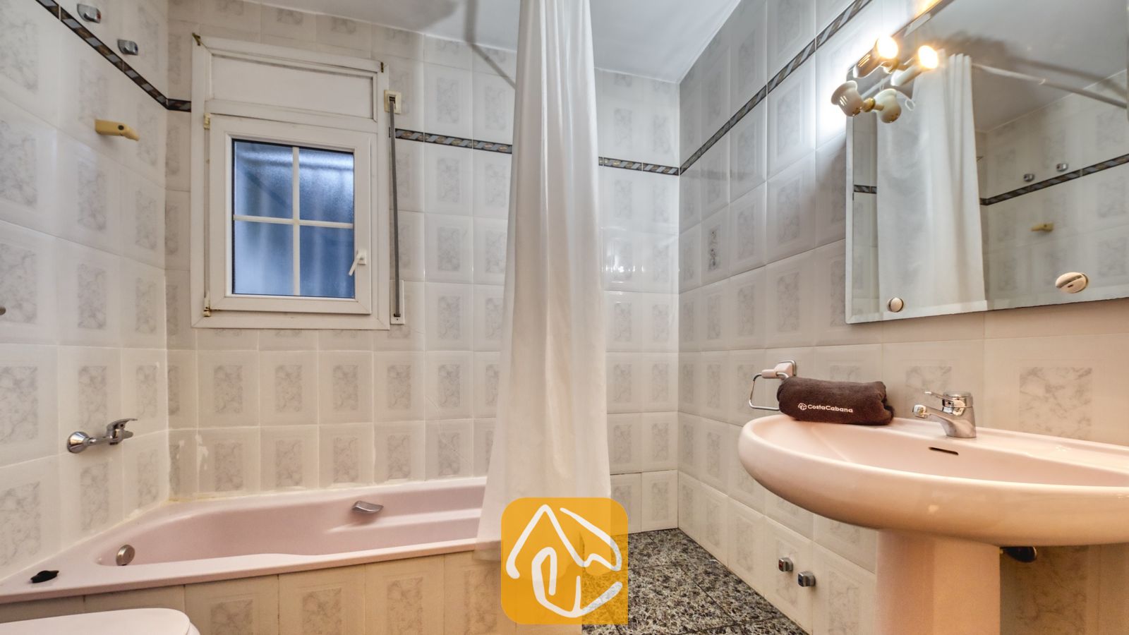 Vakantievilla's Costa Brava Spanje - Villa Rosa - Badkamer met een lichtroze ligbad, bijpassende wastafel op zuil, witte wandtegels met een decoratieve rand en een klein raam met melkglas.