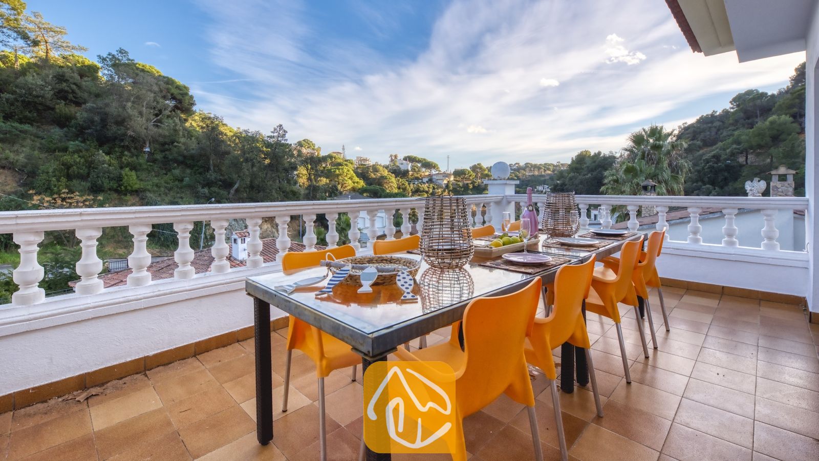 Vakantievilla's Costa Brava Spanje - Villa Rosa - Het balkonterras van Villa Rosa met een glazen eettafel, omringd door oranje plastic stoelen, met uitzicht op beboste heuvels onder een blauwe lucht.