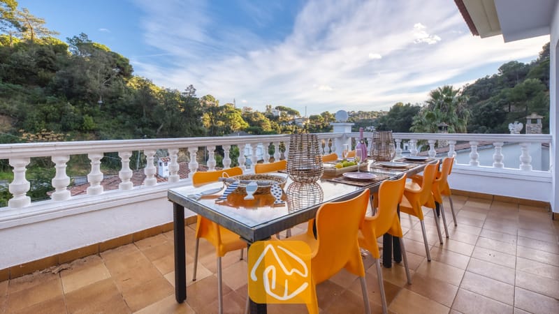 Vakantievilla's Costa Brava Spanje - Villa Rosa - Het balkonterras van Villa Rosa met een glazen eettafel, omringd door oranje plastic stoelen, met uitzicht op beboste heuvels onder een blauwe lucht.