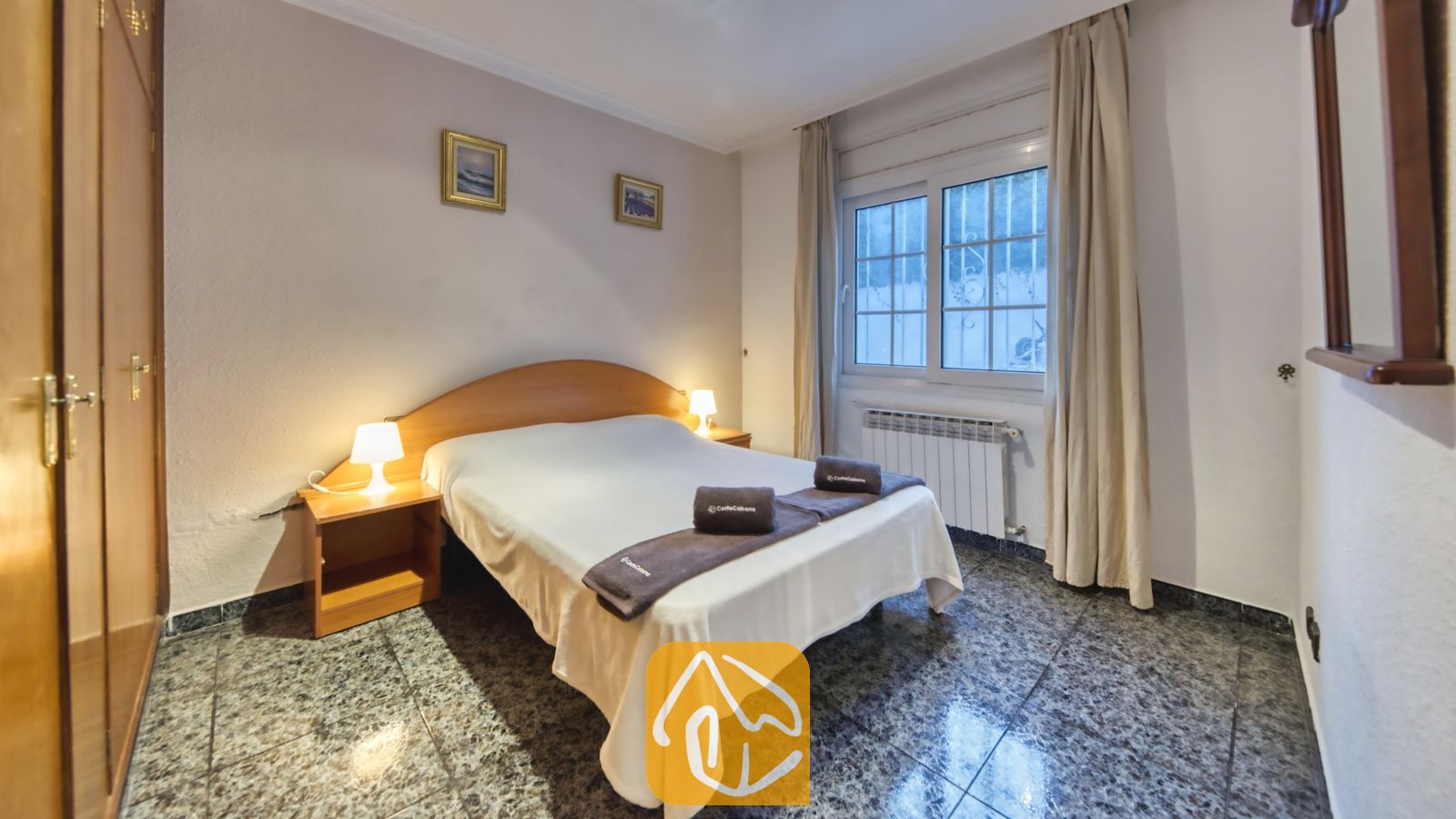 Vakantievilla's Costa Brava Spanje - Villa Rosa - Slaapkamer met een tweepersoonsbed, houten nachtkastjes met lampen, ingelijste kunst aan de muur en een raam met lichte gordijnen.