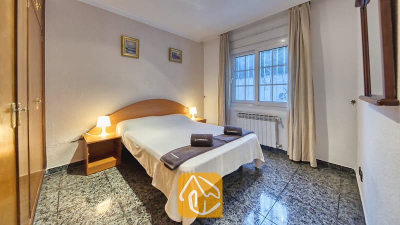 Vakantievilla's Costa Brava Spanje - Villa Rosa - Slaapkamer met een tweepersoonsbed, houten nachtkastjes met lampen, ingelijste kunst aan de muur en een raam met lichte gordijnen.