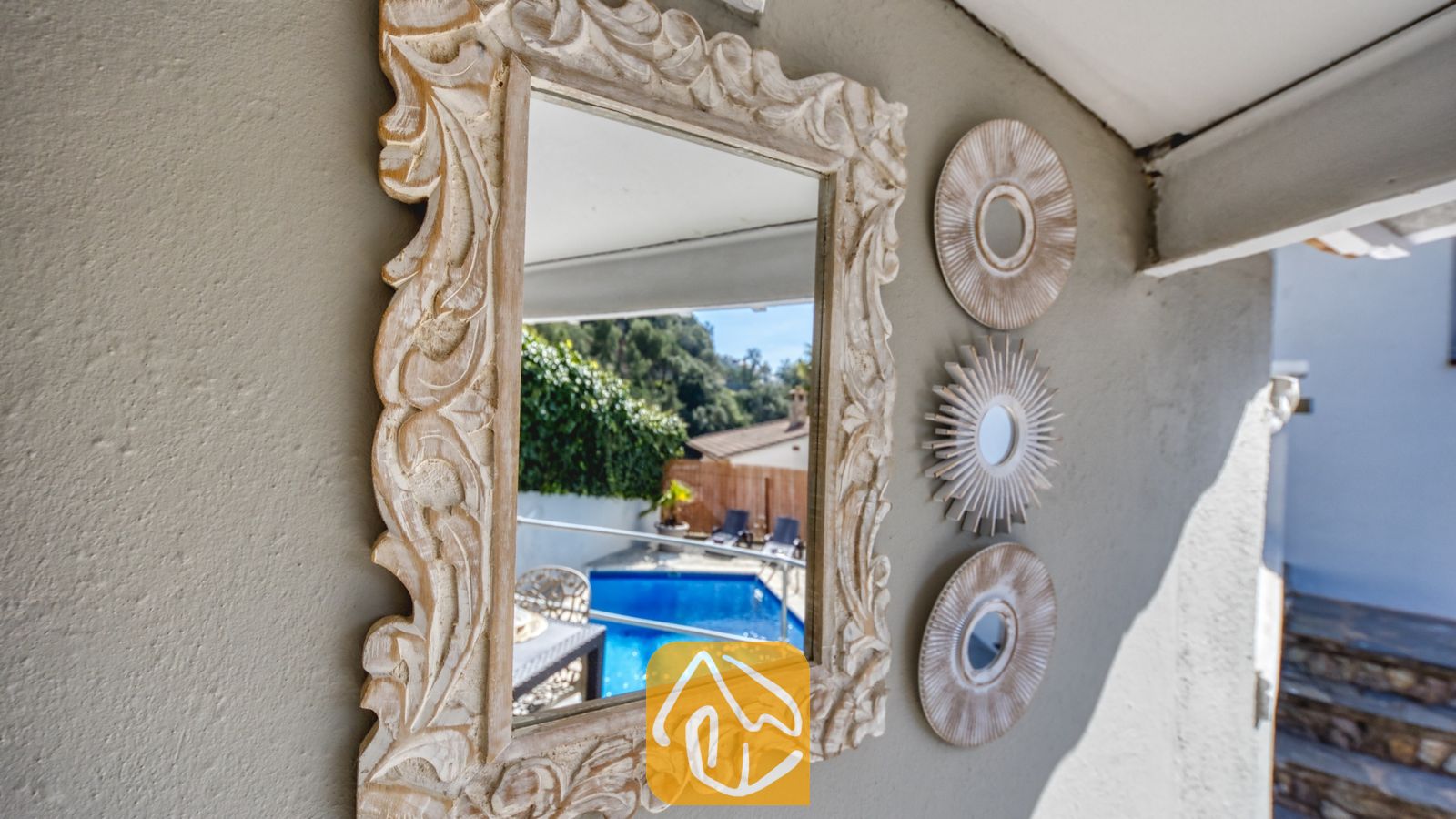 Vakantievilla's Costa Brava Spanje - Villa Rosa - Decoratieve, snijwerk houten spiegel aan een grijze buitenmuur waarin een blauw zwembad en groen worden weerspiegeld bij Villa Rosa.