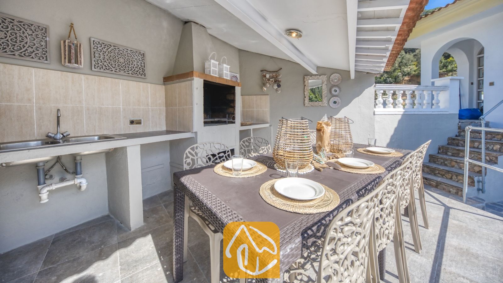 Vakantievilla's Costa Brava Spanje - Villa Rosa - Overdekte eethoek buiten bij Villa Rosa, voorzien van een bruine eettafel met gevlochten placemats, witte borden en bijpassende stoelen, gecombineerd met een ingebouwde barbecue en buitenwastafel.