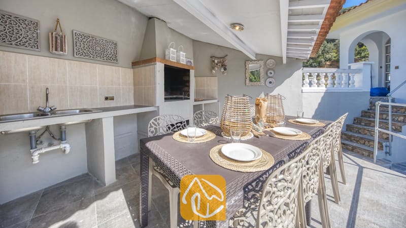 Vakantievilla's Costa Brava Spanje - Villa Rosa - Overdekte eethoek buiten bij Villa Rosa, voorzien van een bruine eettafel met gevlochten placemats, witte borden en bijpassende stoelen, gecombineerd met een ingebouwde barbecue en buitenwastafel.