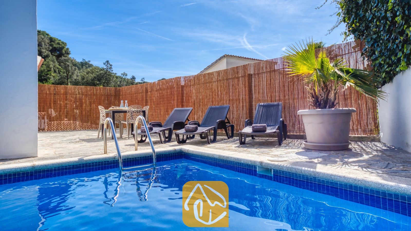 Vakantievilla's Costa Brava Spanje - Villa Rosa - Buitenzwembad met blauwe betegeling en metalen ladder bij Villa Rosa, voorzien van drie donkere ligbedden, een eettafel en een pot met palmboom tegen een rieten schutting.