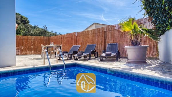 Vakantievilla's Costa Brava Spanje - Villa Rosa - Buitenzwembad met blauwe betegeling en metalen ladder bij Villa Rosa, voorzien van drie donkere ligbedden, een eettafel en een pot met palmboom tegen een rieten schutting.