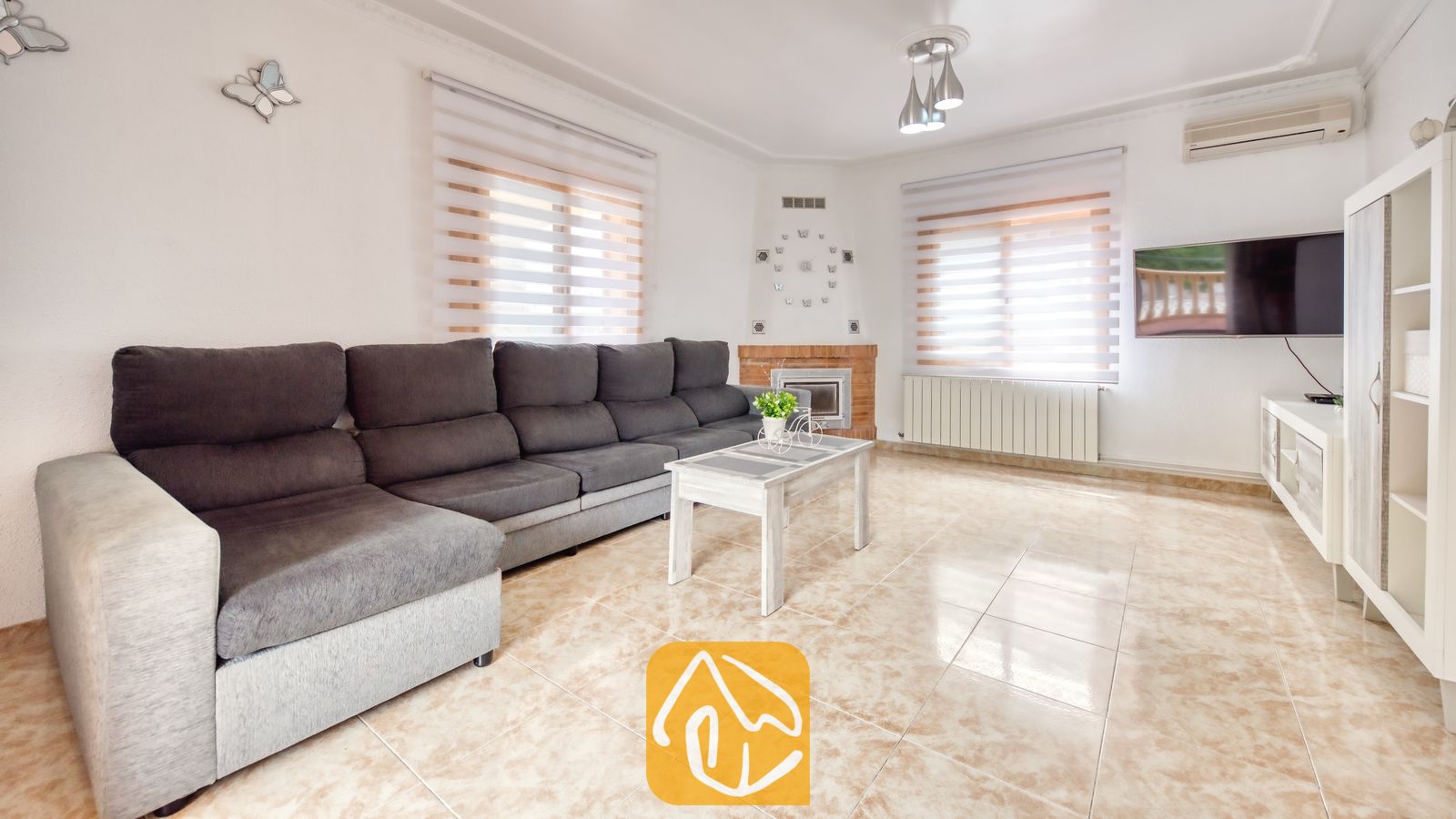 Vakantievilla's Costa Brava Spanje - Villa Sarai - Woonkamer van Villa Sarai met een lange grijze hoekbank, witte tegelvloeren, ramen met zebra-gordijnen en een flatscreen-televisie aan de muur.