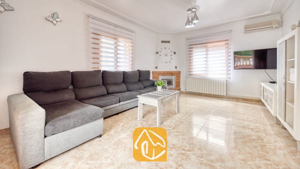 Vakantievilla's Costa Brava Spanje - Villa Sarai - Woonkamer van Villa Sarai met een lange grijze hoekbank, witte tegelvloeren, ramen met zebra-gordijnen en een flatscreen-televisie aan de muur.