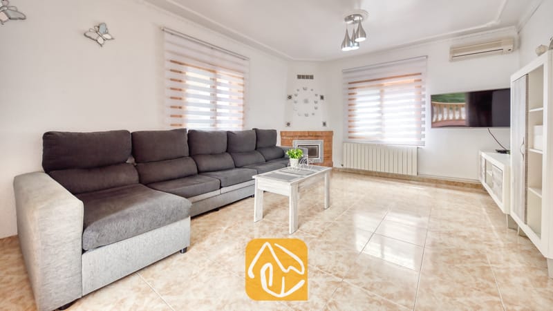 Vakantievilla's Costa Brava Spanje - Villa Sarai - Woonkamer van Villa Sarai met een lange grijze hoekbank, witte tegelvloeren, ramen met zebra-gordijnen en een flatscreen-televisie aan de muur.