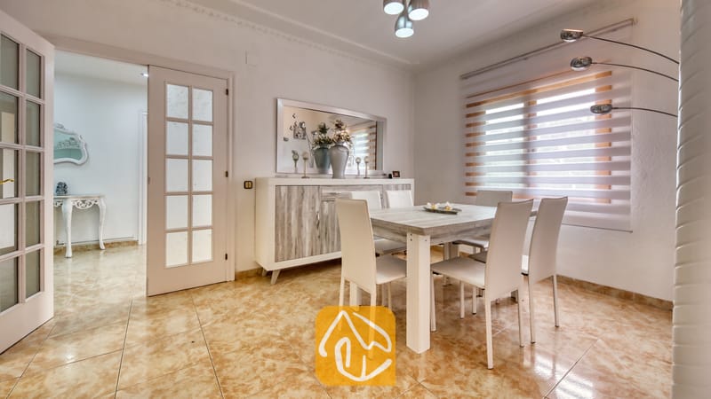Vakantievilla's Costa Brava Spanje - Villa Sarai - Eethoek met een lichthouten tafel, witte gestoffeerde stoelen, een bijpassend dressoir en een raam met gestreepte jaloezieën in Villa Sarai.