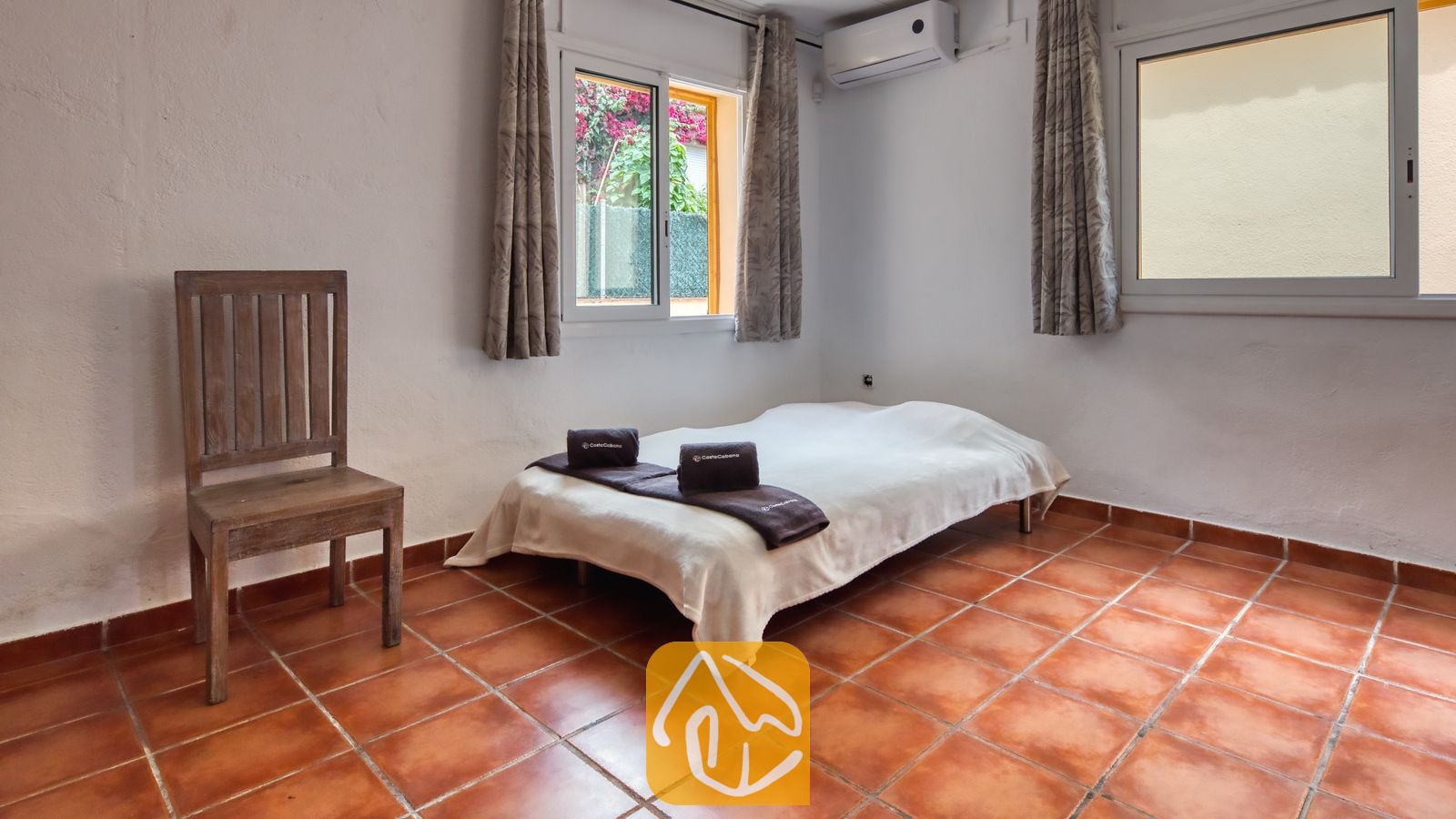 Vakantievilla's Costa Brava Spanje - Villa Sarai - Slaapkamer met een eenpersoonsbed opgemaakt in wit linnen, donker opgevouwen handdoeken, een houten stoel en terracotta plavuizen onder twee ramen.