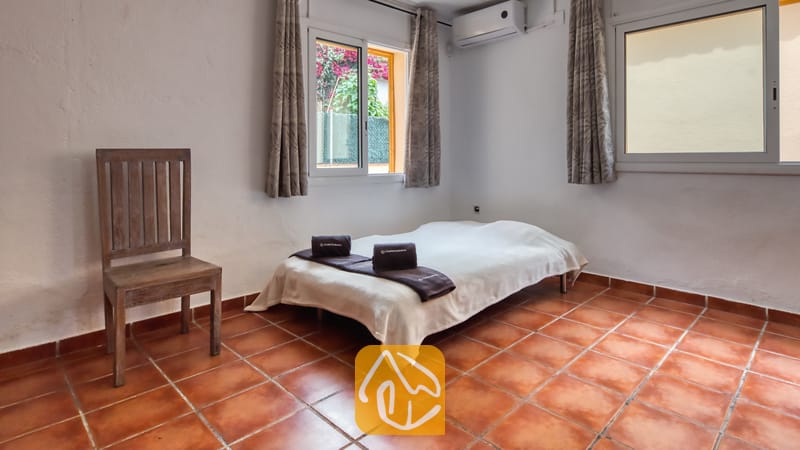 Villas de vacances Costa Brava Espagne - Villa Sarai - Chambre avec un lit simple habillé de linge blanc, des serviettes sombres pliées, une chaise en bois et des sols en terre cuite sous deux fenêtres.