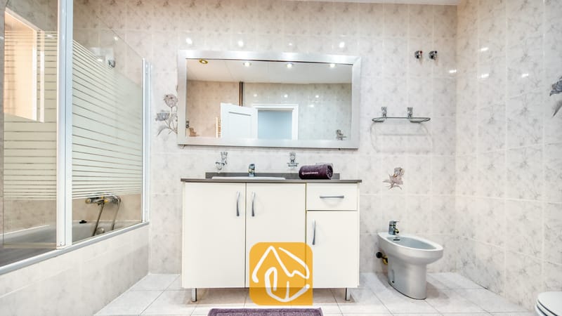 Villas de vacances Costa Brava Espagne - Villa Sarai - Salle de bains comprenant un meuble sous-vasque blanc avec un plan de travail sombre, un grand miroir, un bidet et une baignoire avec pare-douche en verre.