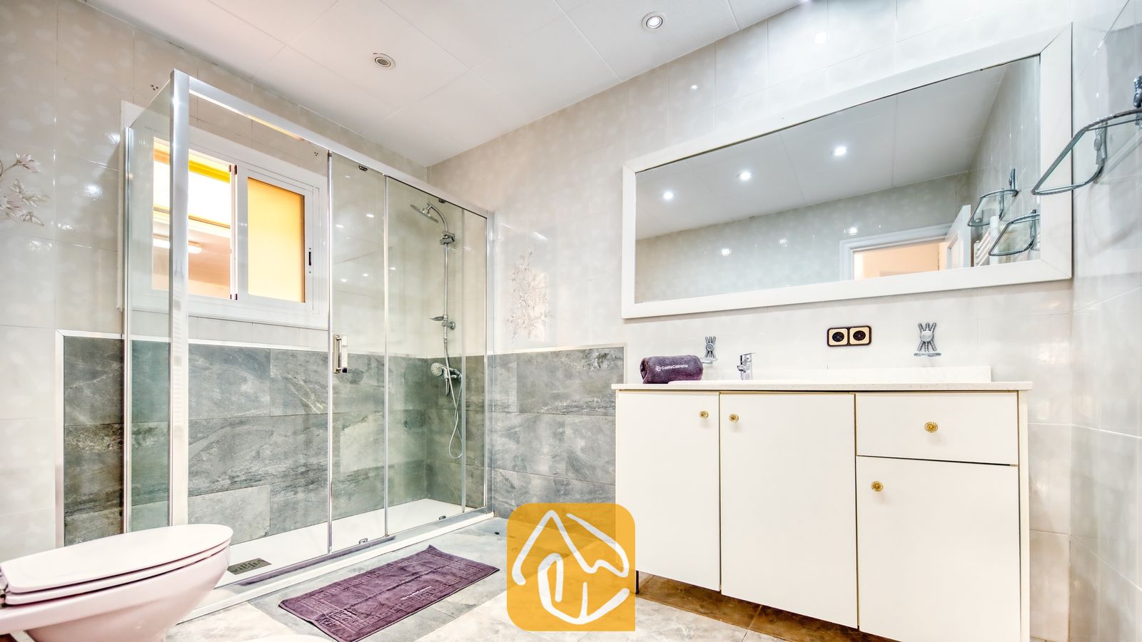 Vakantievilla's Costa Brava Spanje - Villa Sarai - Moderne badkamer met een glazen inloopdouche voorzien van grijs natuursteen, een wit badmeubel met een grote wandspiegel en een toilet.