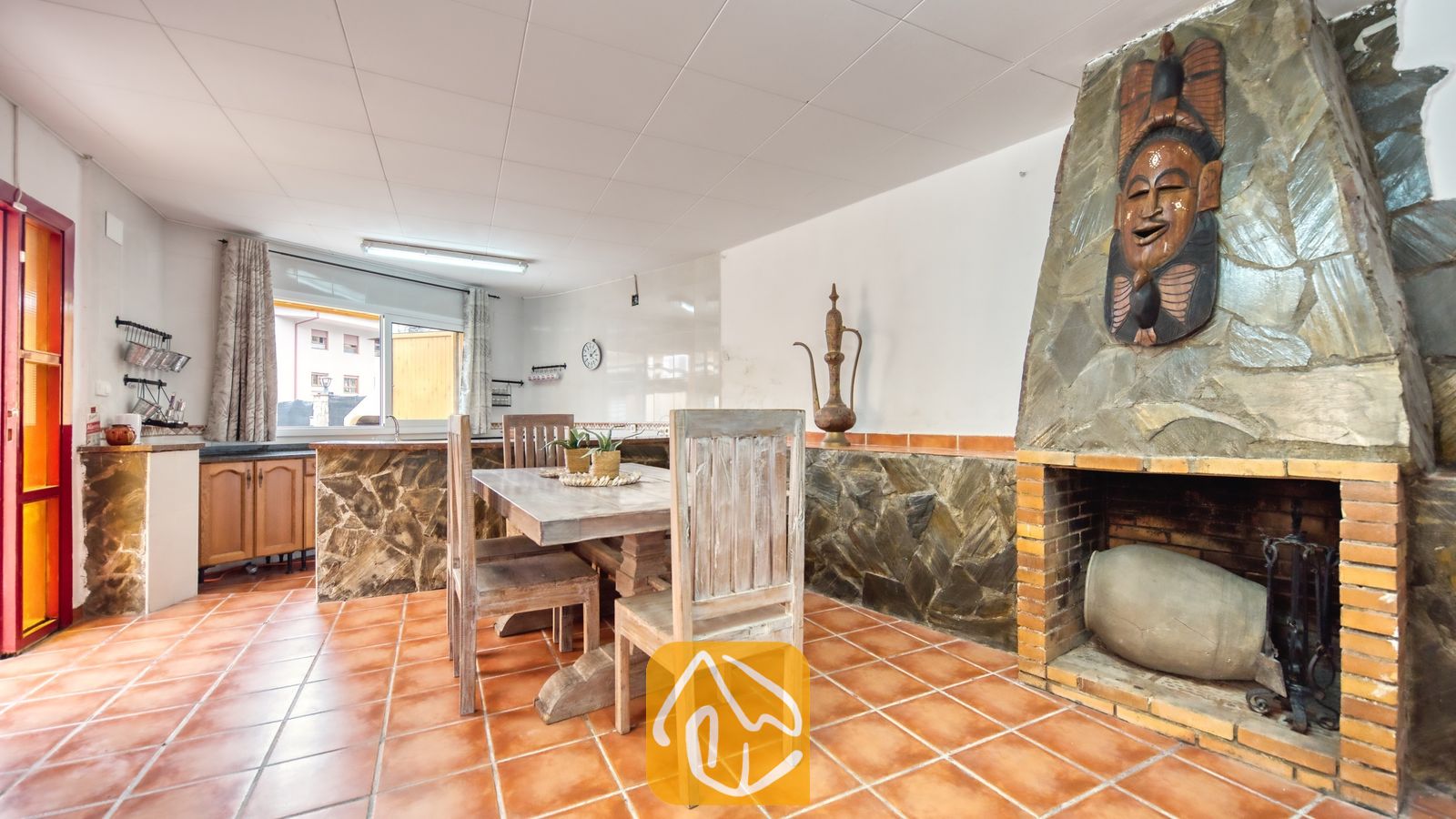 Vakantievilla's Costa Brava Spanje - Villa Sarai - Rustieke eethoek en keuken in Villa Sarai, voorzien van een stenen open haard met een houten masker, verweerde houten stoelen en terracotta plavuizen.