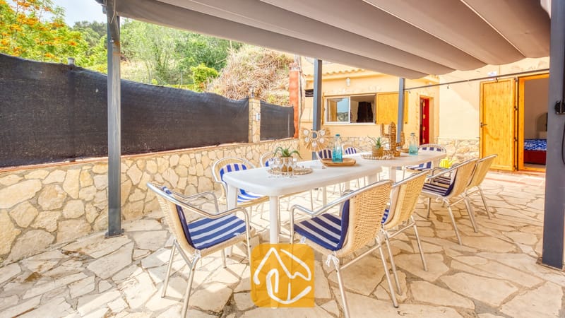 Vakantievilla's Costa Brava Spanje - Villa Sarai - Overdekte eethoek buiten bij Villa Sarai met witte tafels en geweven stoelen voorzien van blauw-witte gestreepte kussens op een stenen terras.
