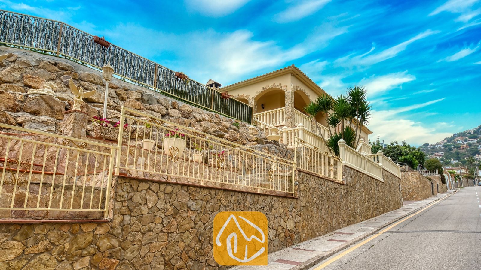 Vakantievilla's Costa Brava Spanje - Villa Sarai - Buitenaanzicht van Villa Sarai met terrasvormige stenen muren in verschillende niveaus, een lichtkleurige railing en palmbomen tegen een blauwe lucht.