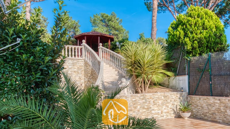 Vakantievilla's Costa Brava Spanje - Villa PrimaDonna - Een stenen buitentrap met balustrades leidt naar een houten prieel bij Villa PrimaDonna, omringd door weelderige, groene palmen en bomen onder een blauwe lucht.