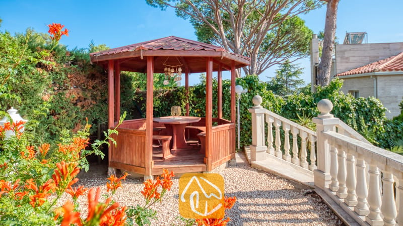 Vakantievilla's Costa Brava Spanje - Villa PrimaDonna - Houten priëel met een tafel en ingebouwde banken in de tuin van Villa PrimaDonna, naast een stenen balustrade trap.