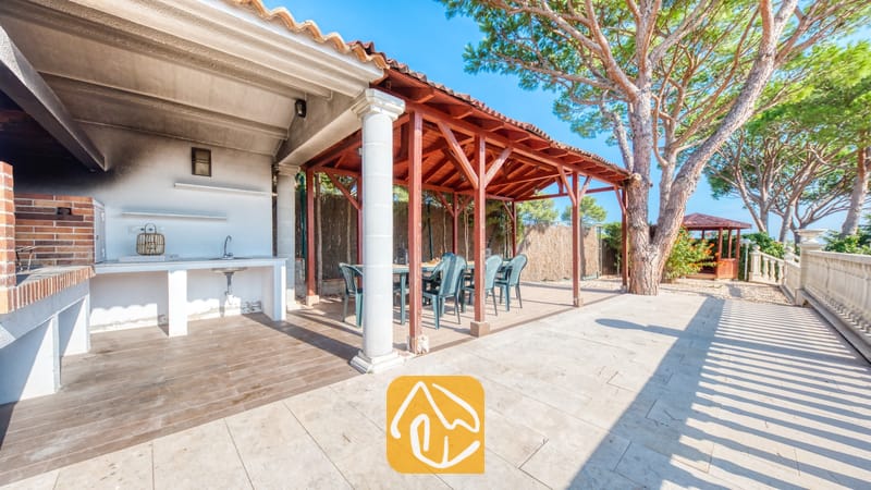Vakantievilla's Costa Brava Spanje - Villa PrimaDonna - Buiteniterras van Villa PrimaDonna met een overdekte eethoek met houten balken, een stenen barbecue-aanrecht met wasbak en omringd door dennenbomen.