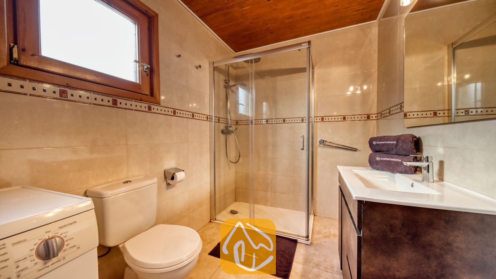 Vakantievilla's Costa Brava Spanje - Villa PrimaDonna - Betegelde badkamer met een glazen douchecabine, een wit toilet, wastafelmeubel met opgestapelde bruine handdoeken, een houten plafond en een wasmachine.