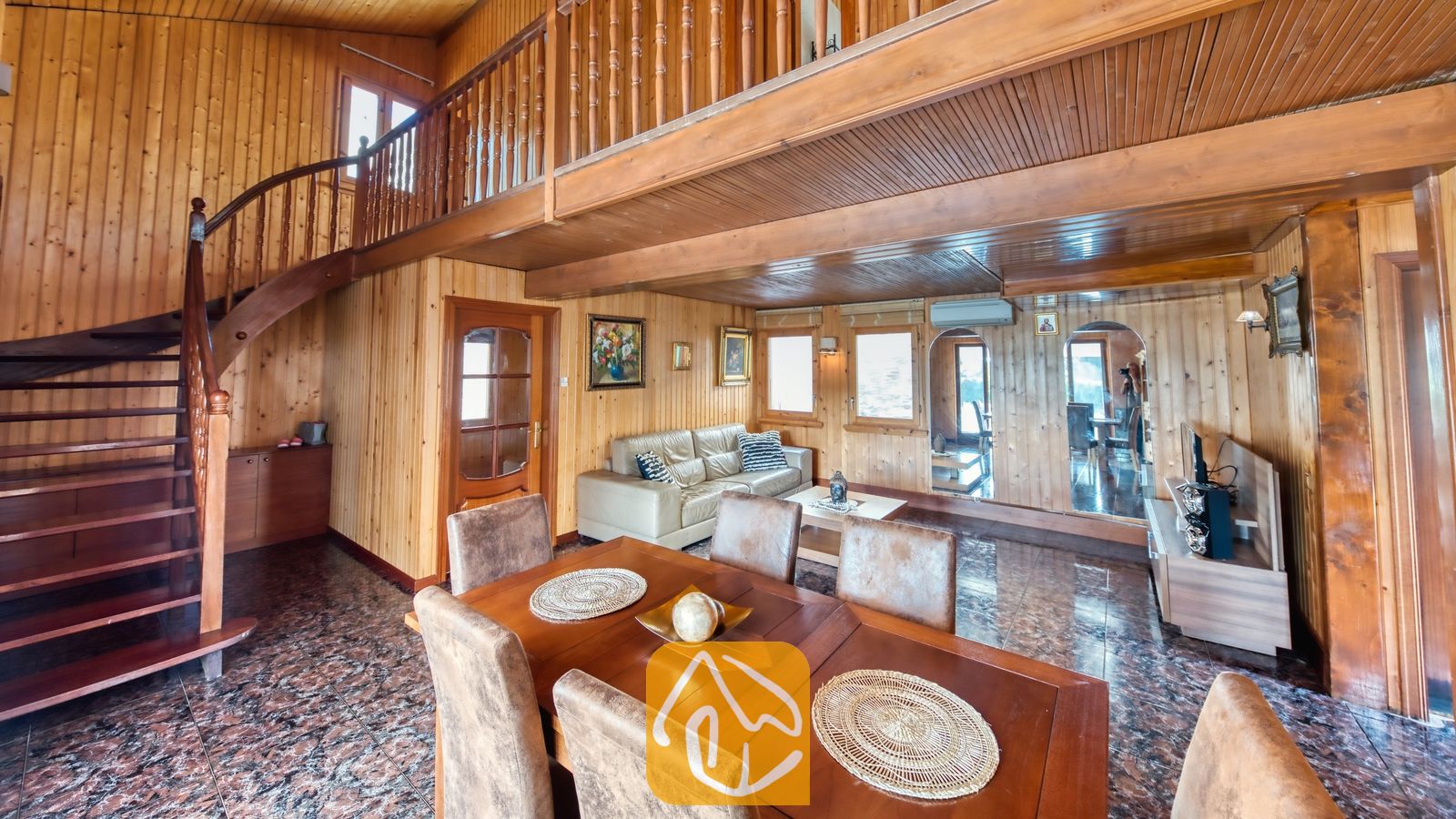 Vakantievilla's Costa Brava Spanje - Villa PrimaDonna - Houten leefgedeelte in Villa PrimaDonna, voorzien van een gebogen trap, een vide met balkon, een eettafel met bruine stoelen, een leren bank en tegelvloeren.