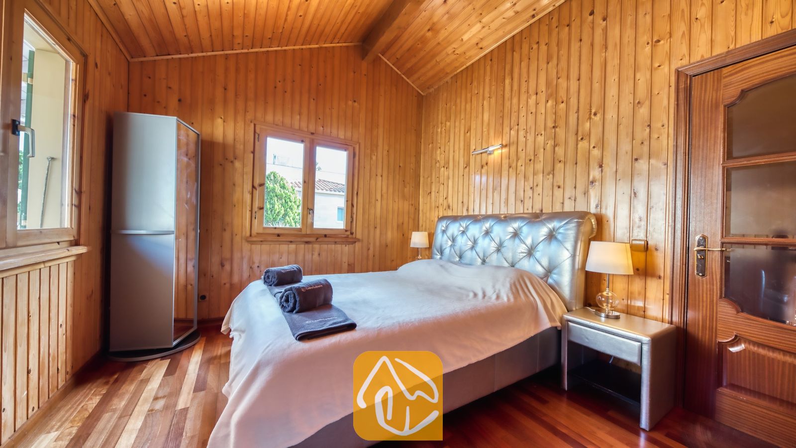 Vakantievilla's Costa Brava Spanje - Villa PrimaDonna - Slaapkamer met houten lambrisering op de muren en het plafond, een bed met een zilverkleurig gecapitonneerd hoofdbord en twee nachtkastjelampjes.