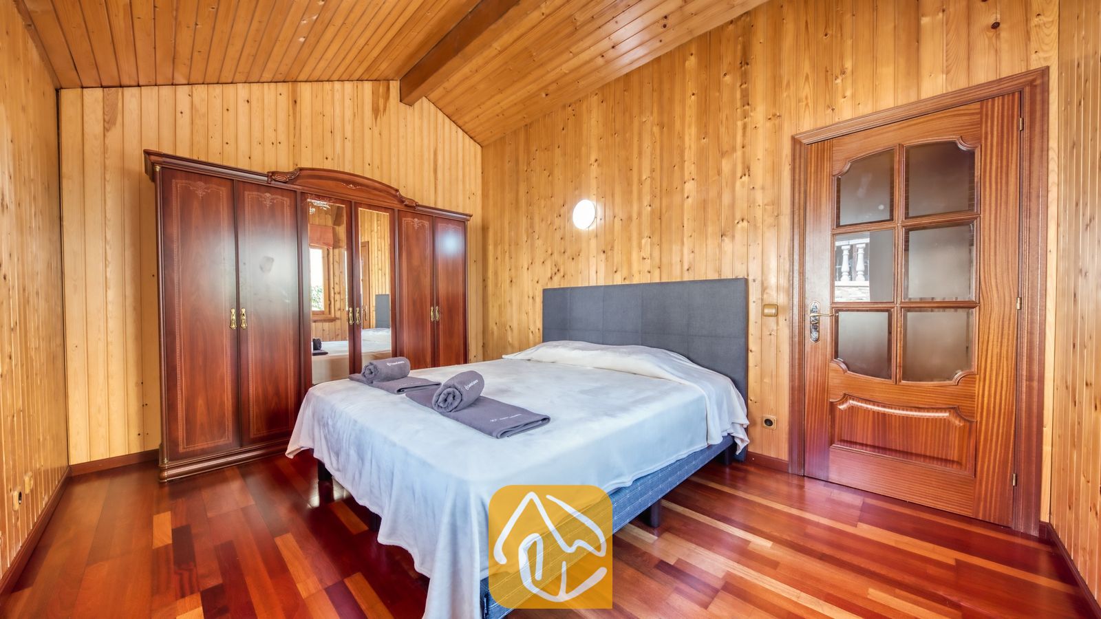 Vakantievilla's Costa Brava Spanje - Villa PrimaDonna - Slaapkamer met houten lambrisering, een tweepersoonsbed met opgerolde grijze handdoeken, een grote donkerhouten kledingkast en een houten deur met glaspanelen.