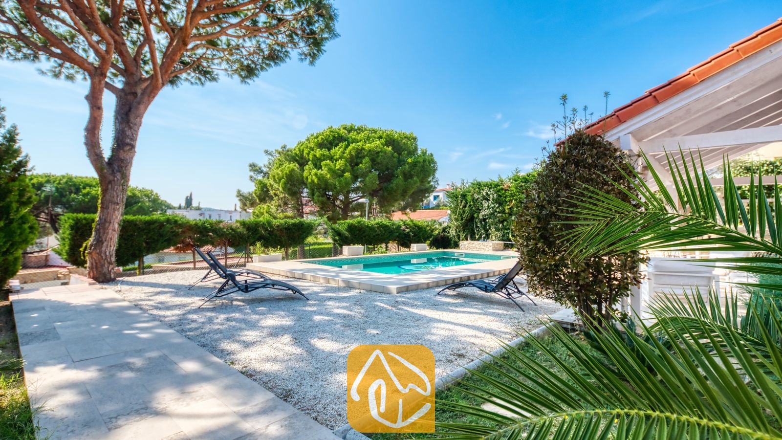 Vakantievilla's Costa Brava Spanje - Villa PrimaDonna - Buitenzwembad bij Villa PrimaDonna met een grindterras, zwarte ligbedden en volwassen dennenbomen onder een blauwe hemel