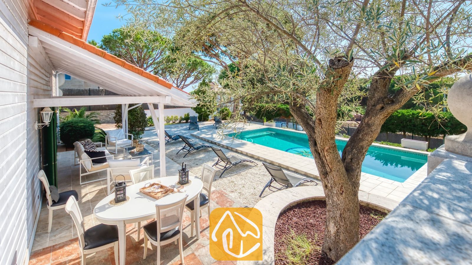 Vakantievilla's Costa Brava Spanje - Villa PrimaDonna - Overdekt buitenterras bij Villa PrimaDonna met een witte ovale tafel, stoelen met zwarte kussens en een zwembadgedeelte omringd door ligstoelen en bomen.