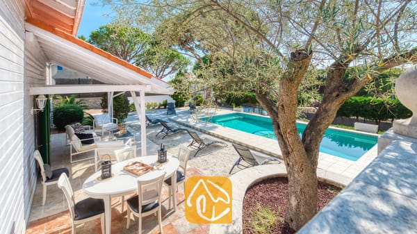 Vakantievilla's Costa Brava Spanje - Villa PrimaDonna - Overdekt buitenterras bij Villa PrimaDonna met een witte ovale tafel, stoelen met zwarte kussens en een zwembadgedeelte omringd door ligstoelen en bomen.