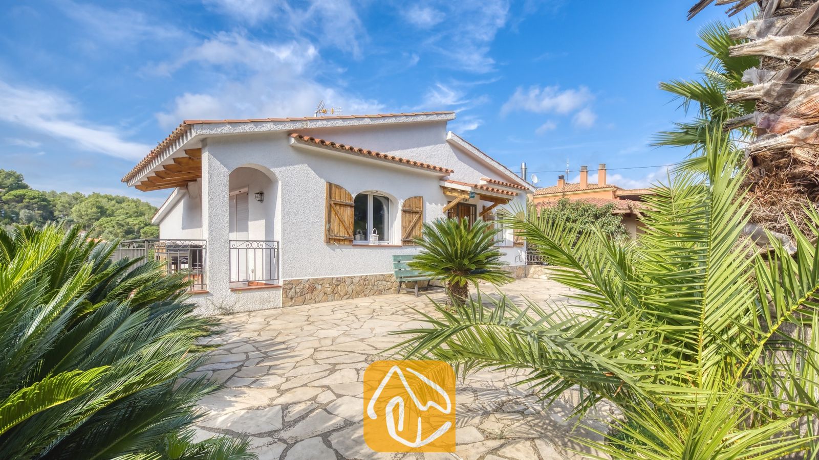 Vakantievilla's Costa Brava Spanje - Villa Nicky - Buitenaanzicht van Villa Nicky met een witpleisterstenen gevel, pannendak, houten luiken, stenen terras en omringende palmbomen onder een blauwe hemel.