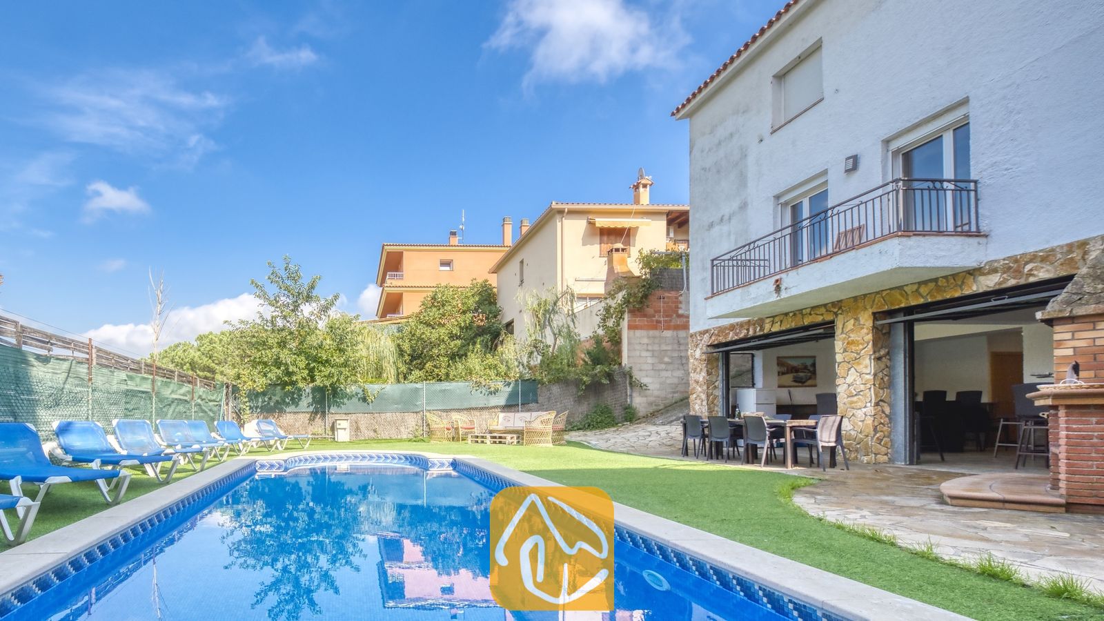 Vakantievilla's Costa Brava Spanje - Villa Nicky - Privézwembad en terras bij Villa Nicky, voorzien van blauwe ligstoelen op kunstgras naast een wit twieverdiepingen tellend landhuis met stenen pilaren en schuifpuien.