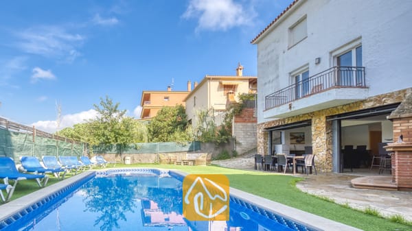 Vakantievilla's Costa Brava Spanje - Villa Nicky - Privézwembad en terras bij Villa Nicky, voorzien van blauwe ligstoelen op kunstgras naast een wit twieverdiepingen tellend landhuis met stenen pilaren en schuifpuien.