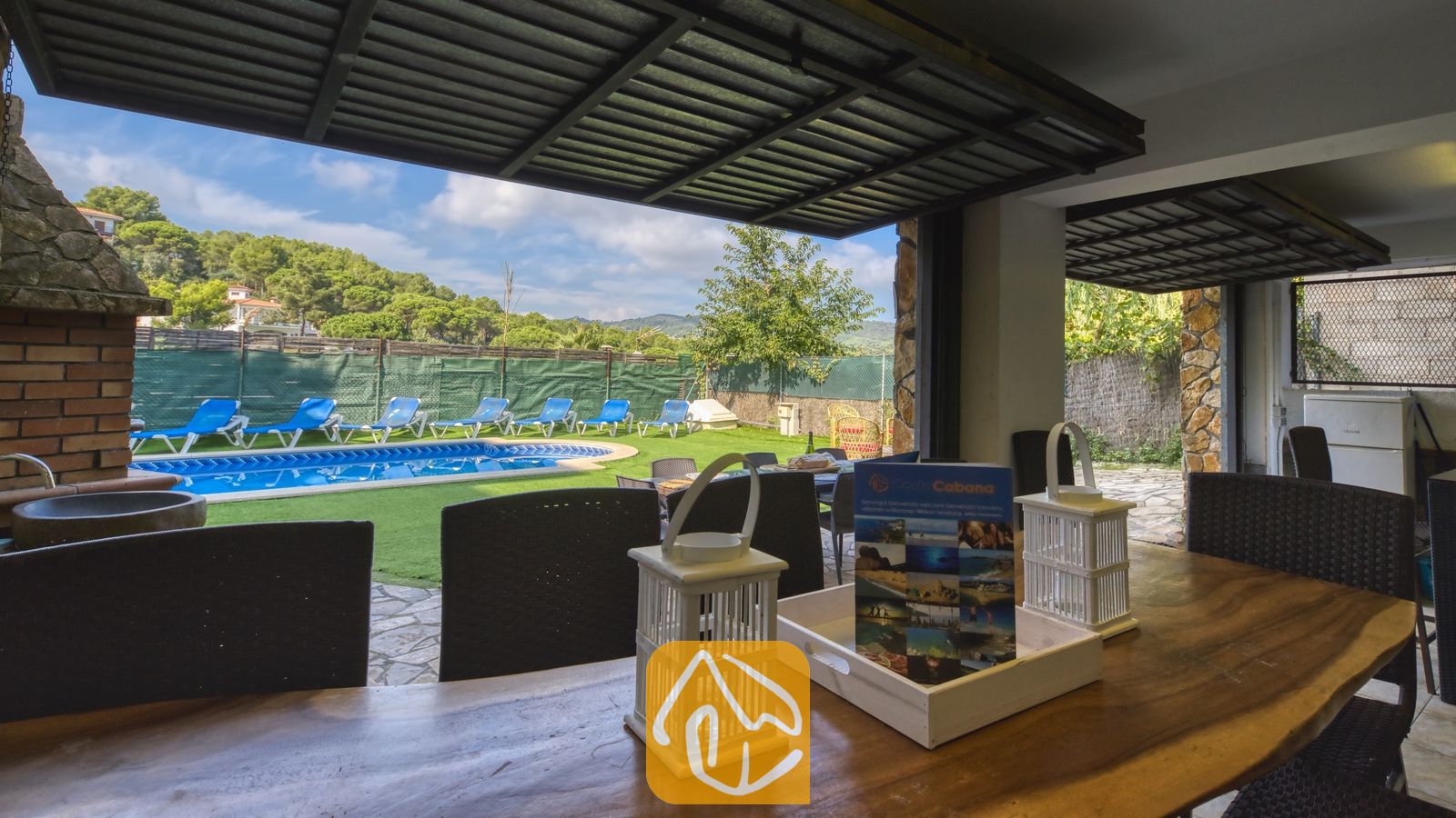Vakantievilla's Costa Brava Spanje - Villa Nicky - Overdekt eethoek buiten bij Villa Nicky met een houten tafel en witte lantaarns, met uitzicht op een zwembad en een groen gazon met ligstoelen.