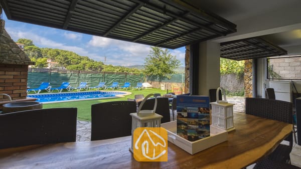 Vakantievilla's Costa Brava Spanje - Villa Nicky - Overdekt eethoek buiten bij Villa Nicky met een houten tafel en witte lantaarns, met uitzicht op een zwembad en een groen gazon met ligstoelen.