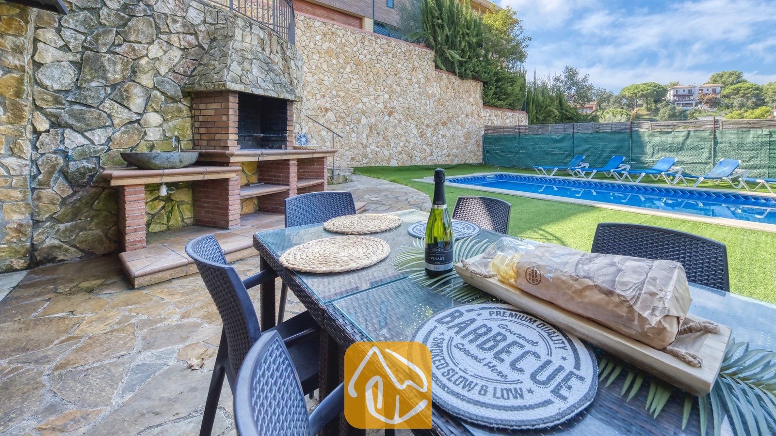 Vakantievilla's Costa Brava Spanje - Villa Nicky - Buiten eettafel gedekt met brood en een fles wijn bij Villa Nicky, voorzien van een ingebouwde stenen barbecue en een zwembad op de achtergrond.