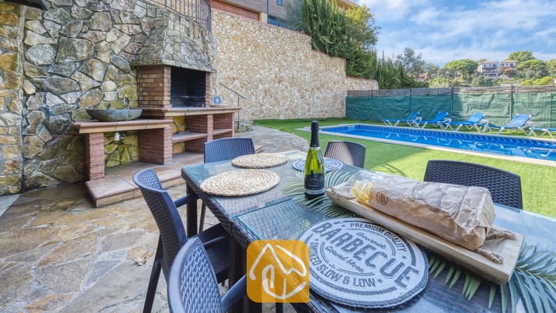 Vakantievilla's Costa Brava Spanje - Villa Nicky - Buiten eettafel gedekt met brood en een fles wijn bij Villa Nicky, voorzien van een ingebouwde stenen barbecue en een zwembad op de achtergrond.