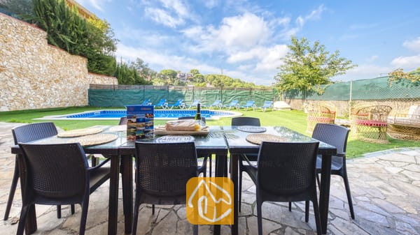 Vakantievilla's Costa Brava Spanje - Villa Nicky - Een eettafel in de buitenlucht op een stenen terras bij Villa Nicky, met uitzicht op een zwembad, een gazon en een blauwe lucht met witte wolken.