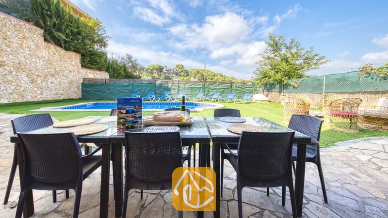 Vakantievilla's Costa Brava Spanje - Villa Nicky - Een eettafel in de buitenlucht op een stenen terras bij Villa Nicky, met uitzicht op een zwembad, een gazon en een blauwe lucht met witte wolken.