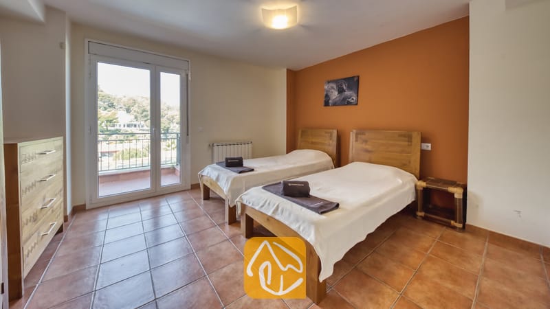 Holiday villas Costa Brava Spain - Villa Nicky - Twin bedroom with wooden headboards, rolled dark towels on white spreads, tiled floors, and a balcony door showing trees outside.