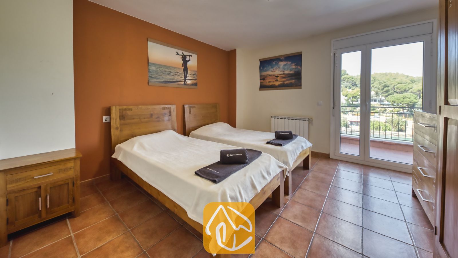 Vakantievilla's Costa Brava Spanje - Villa Nicky - Tweepersoonskamer met twee losse bedden in Villa Nicky, voorzien van twee houten bedden, wit beddengoed, terracotta vloertegels en een balkondeur met uitzicht op groene heuvels.