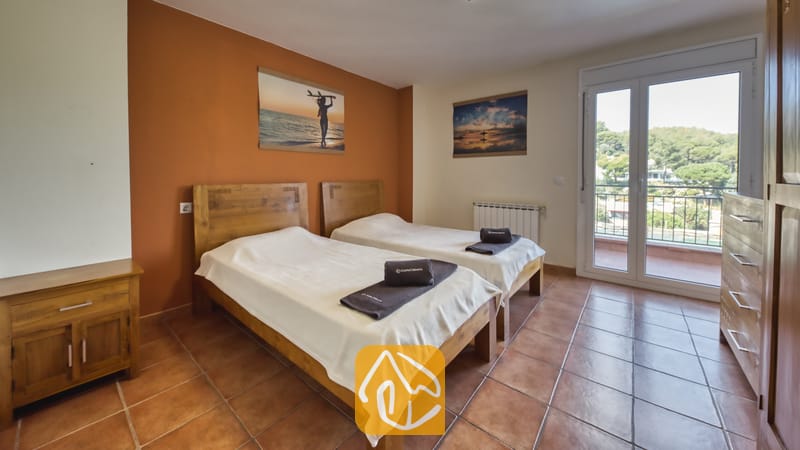 Holiday villas Costa Brava Spain - Villa Nicky - Twin bedroom at Villa Nicky with two wooden beds, white linens, terracotta floor tiles, and a balcony door showing green hills.
