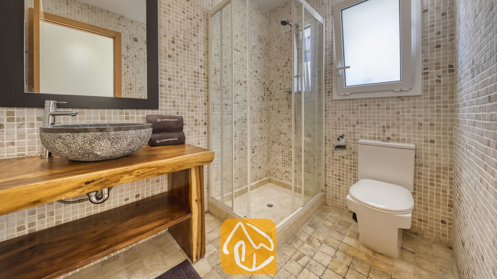 Vakantievilla's Costa Brava Spanje - Villa Nicky - Moderne badkamer met een glazen hoekdouche, een stenen waskom op een houten wastafelmeubel met natuurlijke boomrand, en muren van beige mozaïektegels.