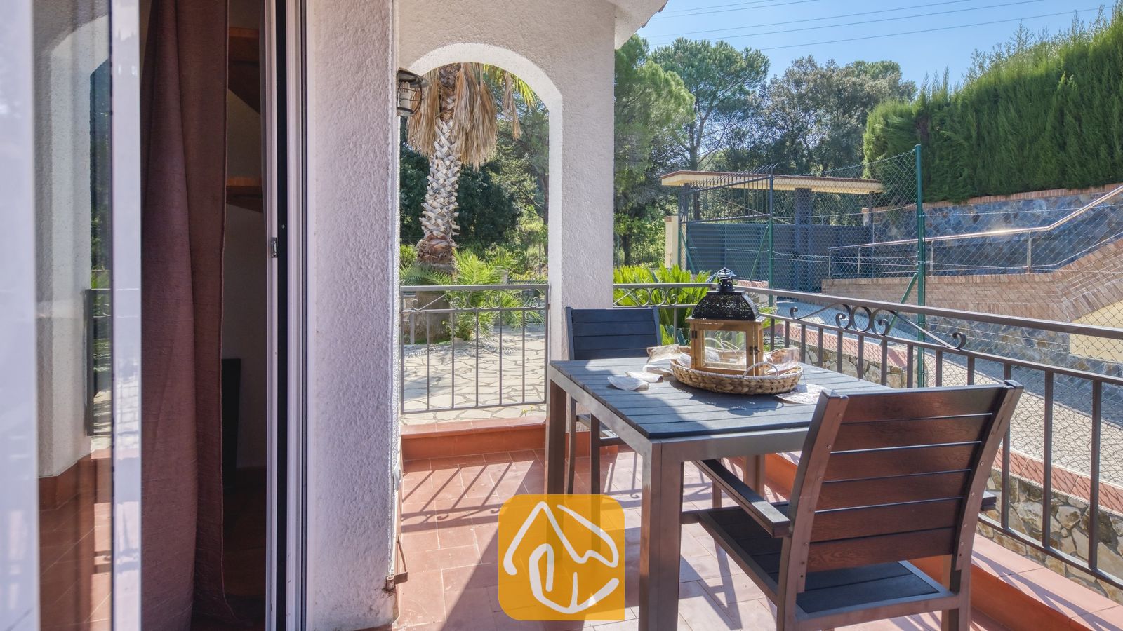 Vakantievilla's Costa Brava Spanje - Villa Nicky - Balkon van Villa Nicky met een donkerhouten eettafel, stoelen, een decoratieve lantaarn en tegelvloer met uitzicht op bomen en omheiningen.