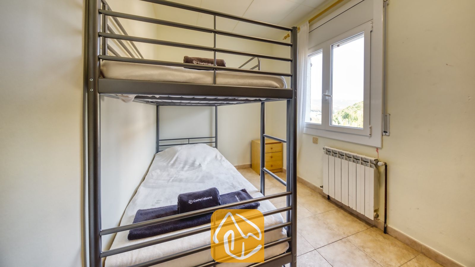 Vakantievilla's Costa Brava Spanje - Villa Nicky - Slaapkamer met grijsmetalen stapelbed, witte beddengoed, opgevouwen donkere handdoeken, een klein houten nachtkastje en een raam met uitzicht naar buiten.