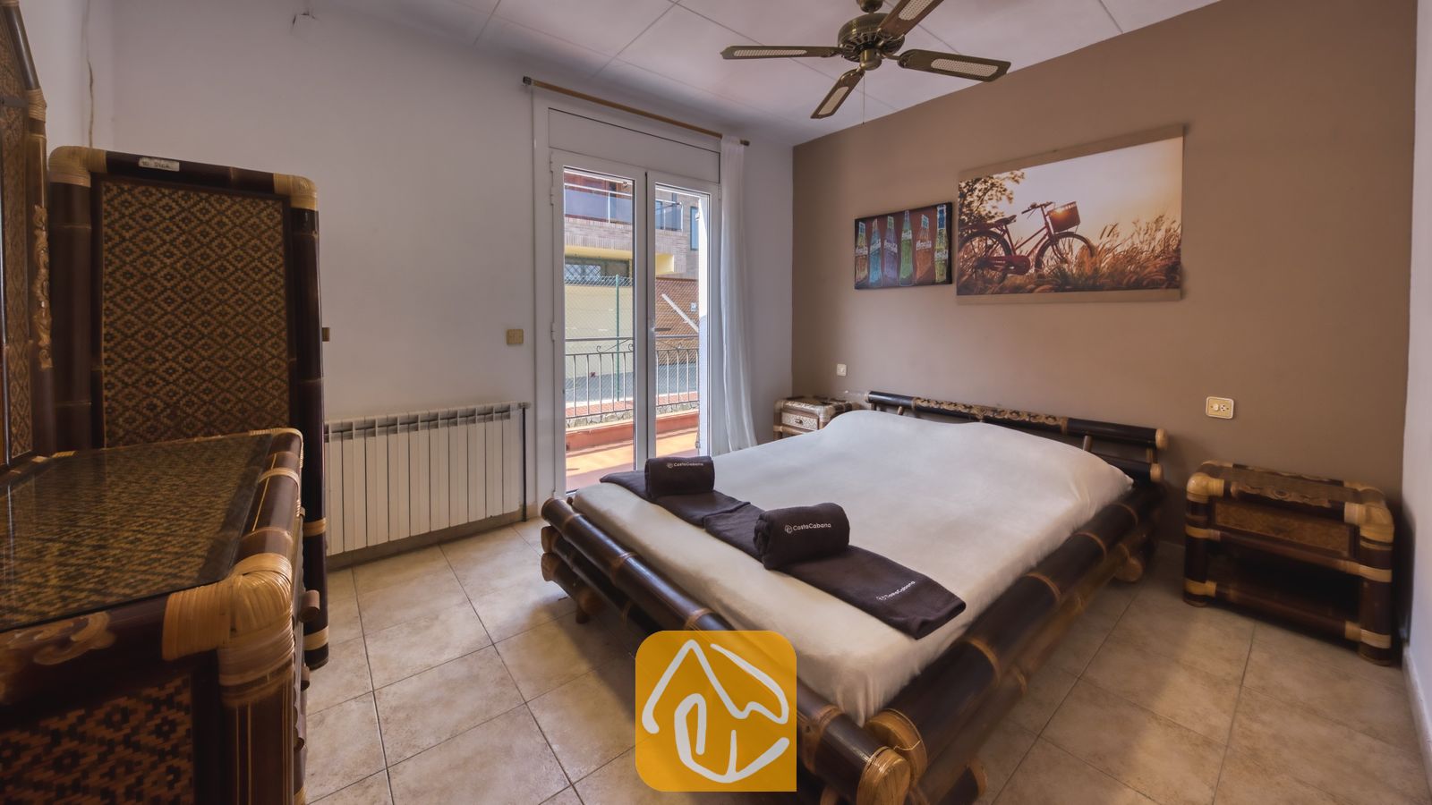 Vakantievilla's Costa Brava Spanje - Villa Nicky - Slaapkamer voorzien van een bamboe bedframe met opgerolde bruine handdoeken, tegelvloeren, bijpassend donker houten meubilair en glazen deuren die uitkomen op een terras.