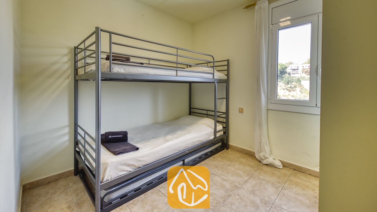 Vakantievilla's Costa Brava Spanje - Villa Nicky - Slaapkamer voorzien van een grijs metalen stapelbed met wit beddengoed en opgevouwen donkere handdoeken, een tegelvloer en een klein raam met witte gordijnen.