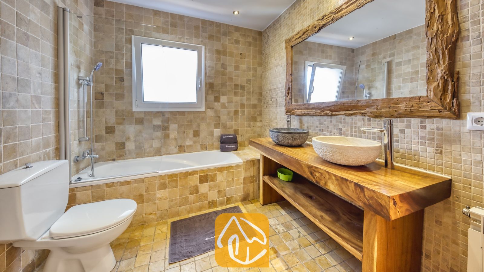 Vakantievilla's Costa Brava Spanje - Villa Nicky - Badkamer met beige betegelde muren, een ligbad met douchekraan, een houten badkamermeubel met twee stenen wastafels en een grote spiegel met een rustiek houten frame.