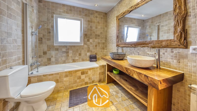 Holiday villas Costa Brava Spain - Villa Nicky - Bathroom with beige tiled walls, a bathtub with shower fixture, a wooden vanity featuring two stone sinks, and a large mirror with a rustic wooden frame.